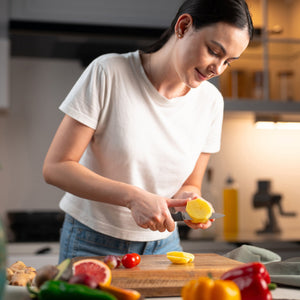 A woman in a white t-shirt smiles while slicing a yellow fruit with the Zyliss Paring Knife Serrated 10cm/4, featuring an ergonomic handle, surrounded by tomatoes, peppers, and grapefruit on a wooden cutting board. 
