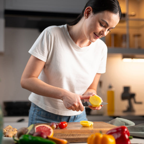 A woman in a white t-shirt smiles while slicing a yellow fruit with the Zyliss Paring Knife Serrated 10cm/4, featuring an ergonomic handle, surrounded by tomatoes, peppers, and grapefruit on a wooden cutting board. 