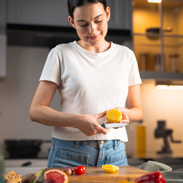 In a kitchen, a woman in a white t-shirt smiles as she squeezes lemon over fruits and veggies, using the Zyliss Paring Knife Serrated 10cm / 4 with an ergonomic, eco-friendly handle on a wooden cutting board. 