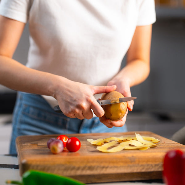 A person in a white shirt and jeans peels a potato with the Zyliss Paring Knife 9cm / 3½ (eco-friendly handle) over a wooden cutting board covered in potato peels, cherry tomatoes, and shallots. 