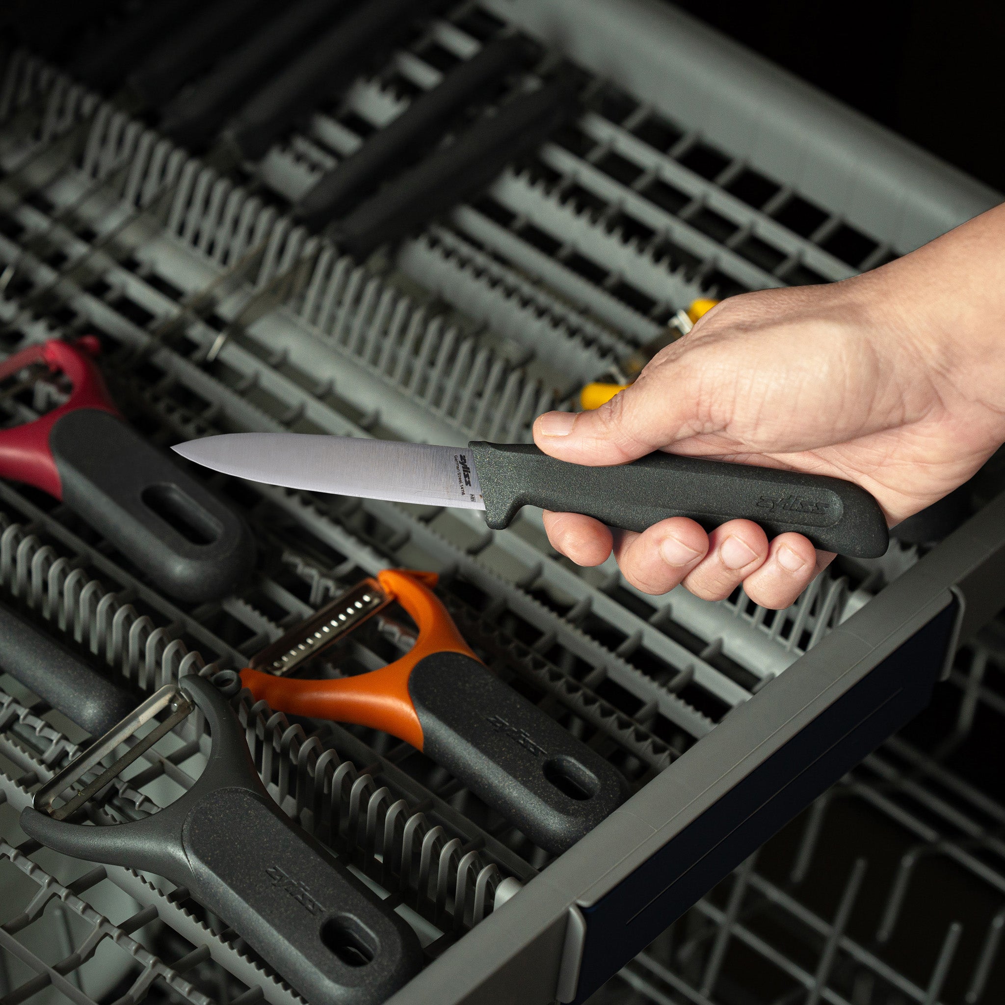 A hand holds a paring knife with a black handle above an open dishwasher rack containing several vegetable peelers with different colored handles.