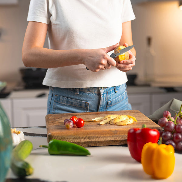 Wearing a white shirt and jeans, a person uses the Zyliss Paring Knife 9cm / 3½ with an eco-friendly handle to slice a potato on a wooden cutting board in a kitchen amid fresh vegetables and grapes. 