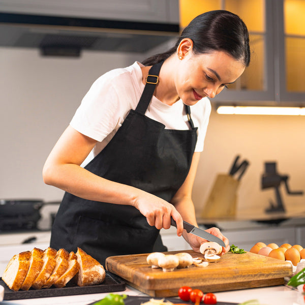 A woman in a black apron slices mushrooms with the Zyliss Vegetable Knife 9cm / 3½ on a wooden cutting board in a modern kitchen, surrounded by bread, eggs, and vegetables. 