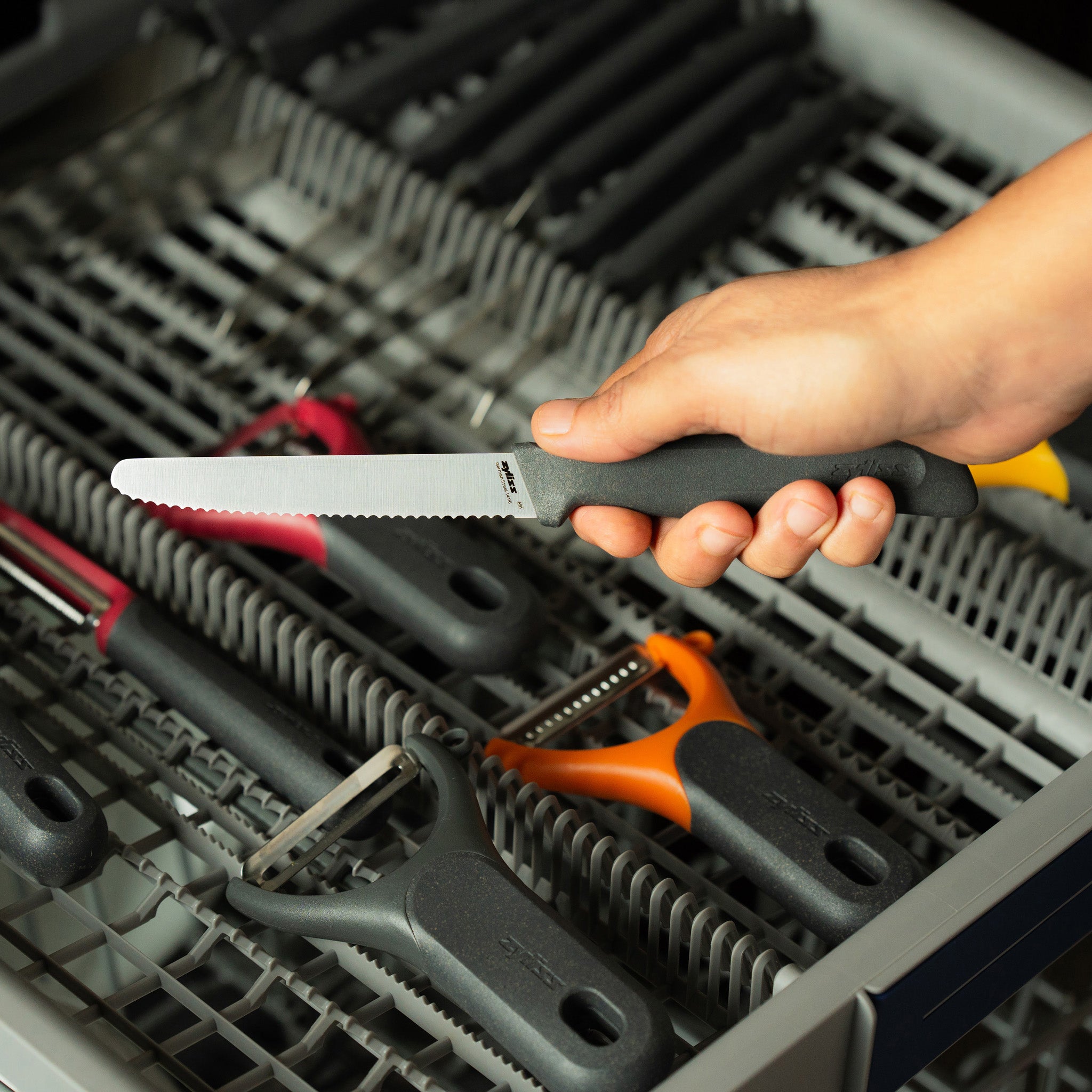 A hand holds a serrated kitchen knife over an open dishwasher drawer filled with various utensils, including peelers and knives.
