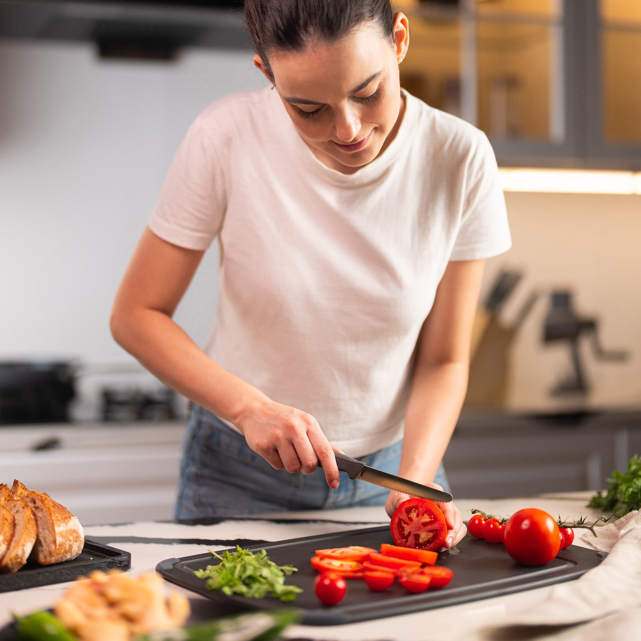A woman in a white t-shirt slices tomatoes on a black cutting board in a modern kitchen using the Zyliss Tomato Knife Serrated 11cm / 4½, which has an eco-friendly handle. Fresh vegetables and bread are spread out on the counter around her. 