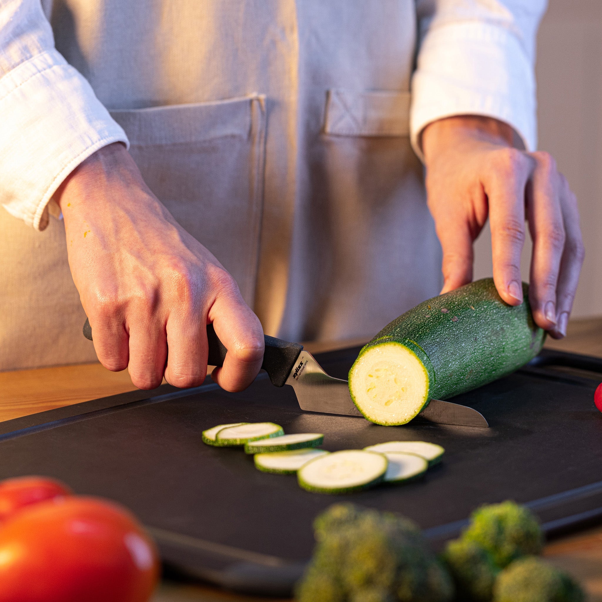Wearing a beige apron, a person uses the Zyliss Offset Knife 10cm / 4 for precision slicing, cutting zucchini on a board. Slices of zucchini rest on the board, with tomatoes and broccoli visible nearby. 