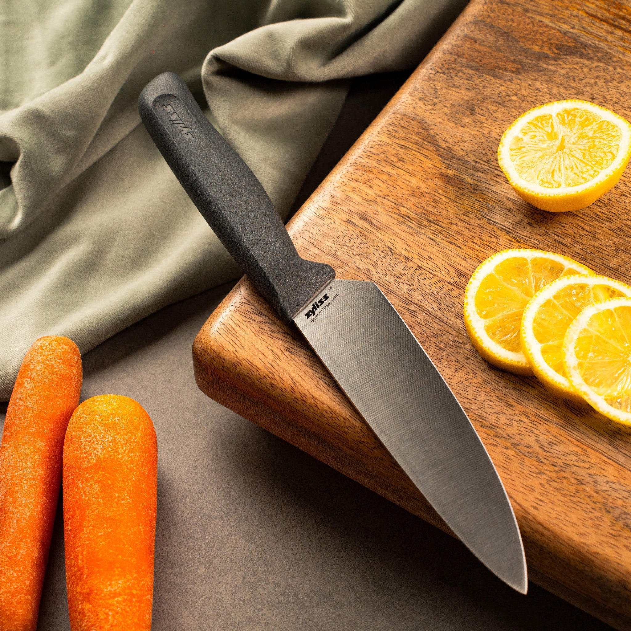 A kitchen knife with a black handle rests on a wooden cutting board, next to slices of lemon. Two whole carrots and a light green cloth are nearby. The scene is well-lit and arranged for food preparation.