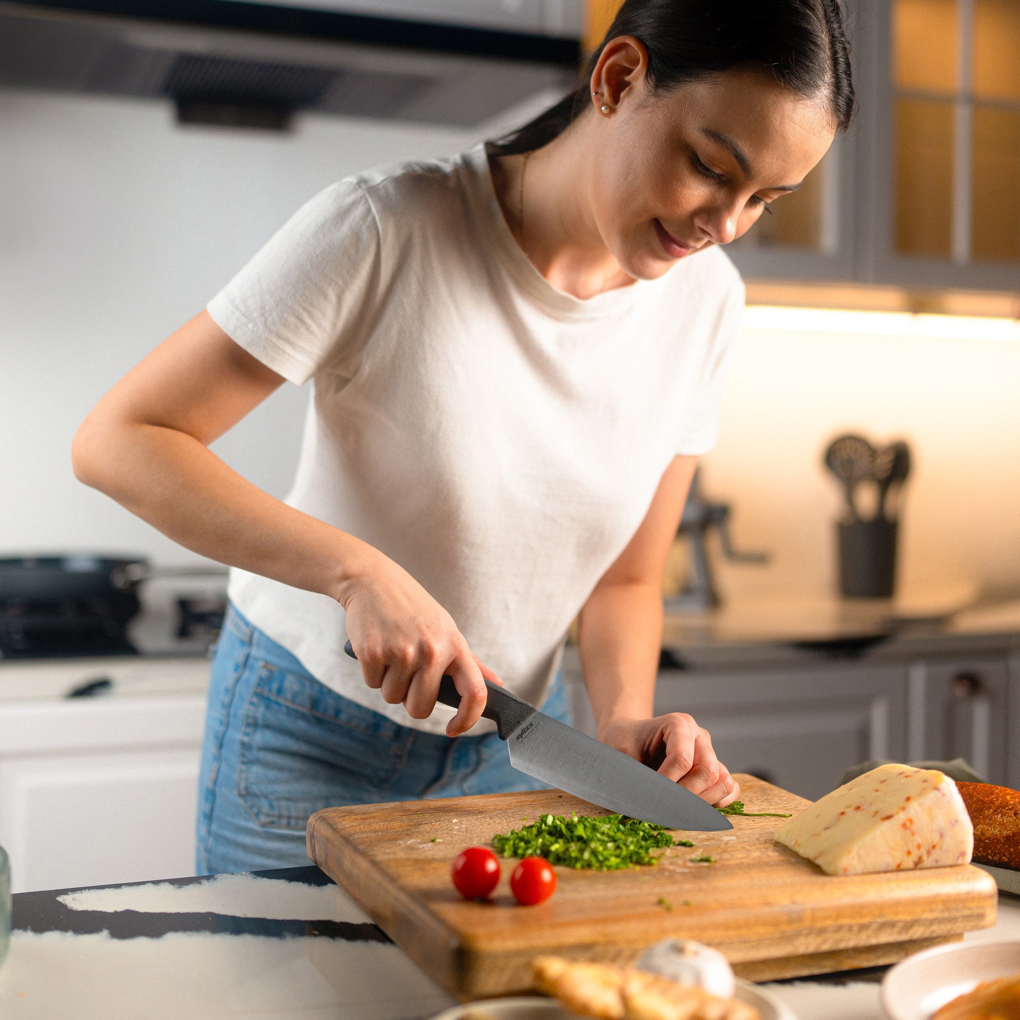 A woman in a white t-shirt chops green herbs on a wooden cutting board in a modern kitchen, with cheese and tomatoes nearby.
