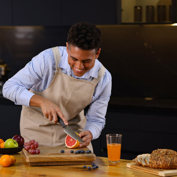 #15cm/6"
A smiling person in an apron uses the Zyliss Chef Knife 15cm / 6 with an ergonomic handle to slice a grapefruit on a wooden cutting board in a sustainable kitchen, surrounded by grapes, apples, blueberries, juice, and sliced bread.