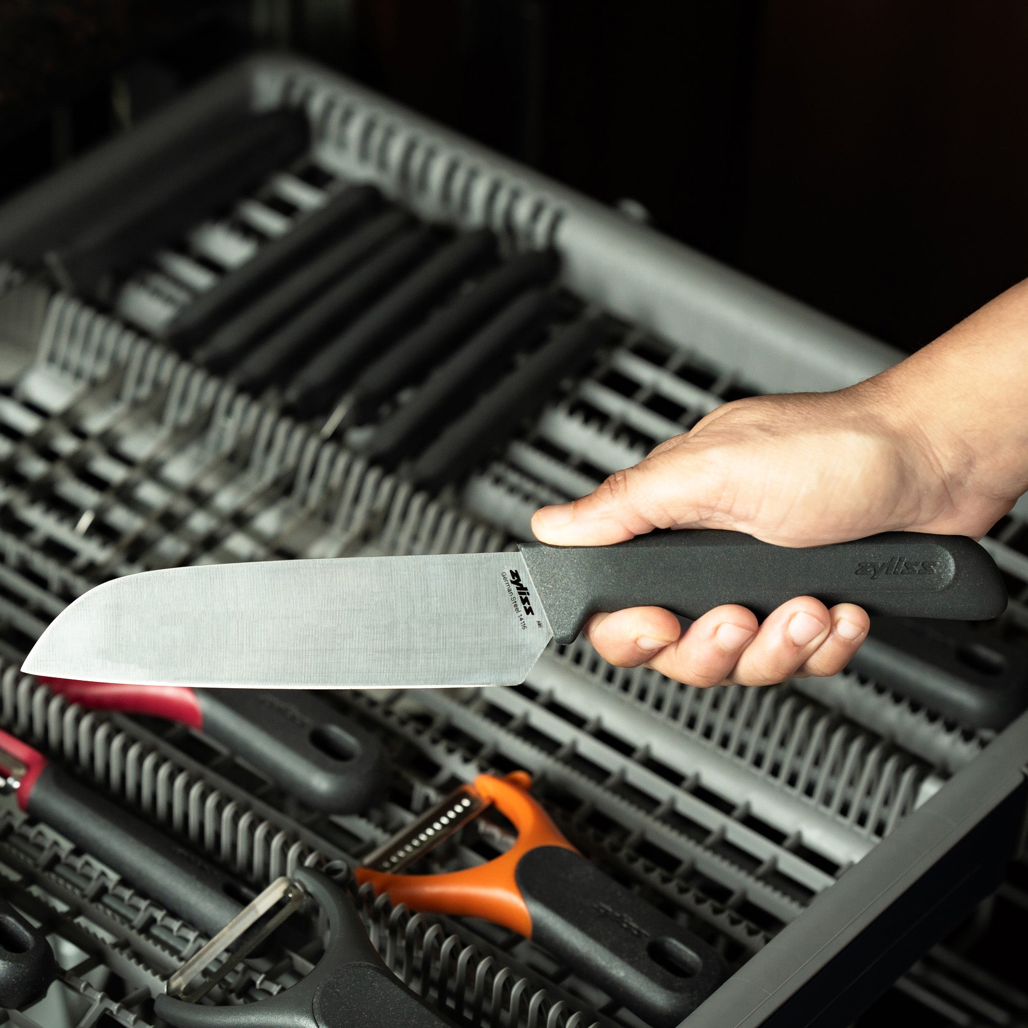 A hand holding a large kitchen knife with a black handle over an open dishwasher filled with utensils and kitchen tools.