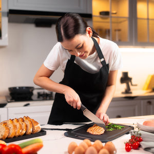 #15cm/6"
In a modern kitchen, a woman in a black apron uses the Zyliss Santoku Knife to slice grilled chicken on a cutting board, surrounded by bread, eggs, and vegetables.