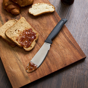 A slice of toasted bread with jam and a Zyliss Spreading Knife 12cm/5" rests on a wooden cutting board, next to more toast and a croissant, set on a dark wooden table.