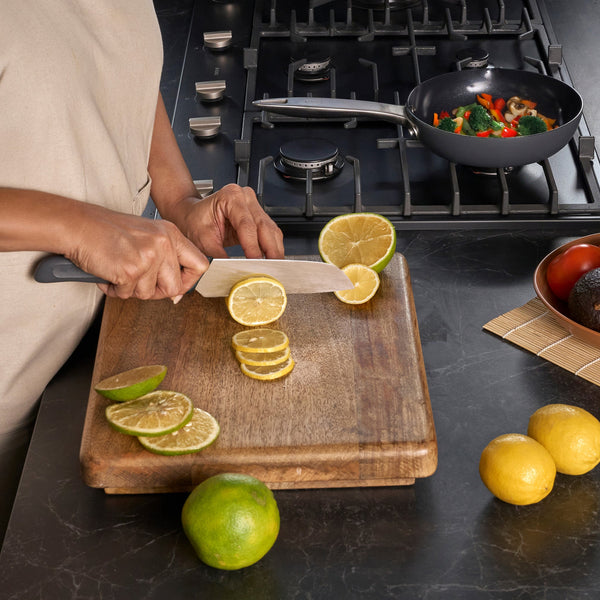 #17cm/7"
A person uses a Zyliss Santoku Knife with an ergonomic handle to slice a lemon on a wooden cutting board next to limes and whole lemons, while vegetables cook in a pan on the stove in the background.