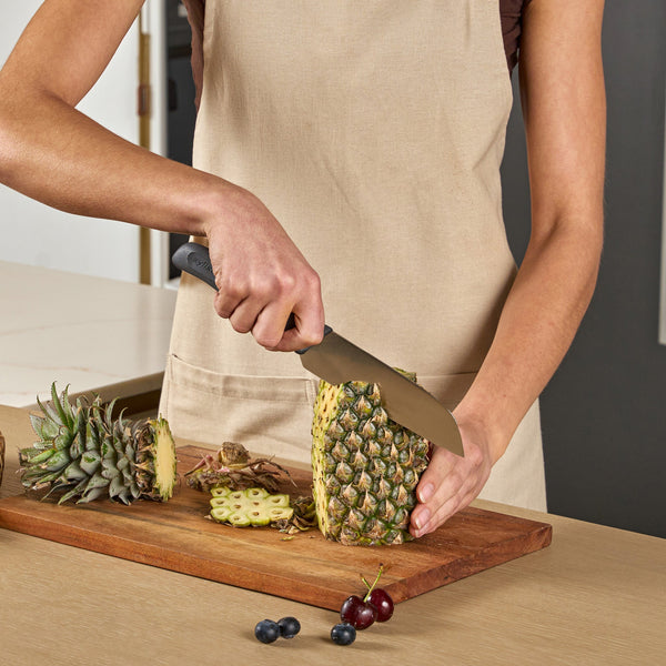 #17cm/7"
Wearing a beige apron, a person uses a Zyliss Santoku Knife with an ergonomic handle to slice a pineapple on a wooden cutting board, surrounded by pineapple tops, blueberries, and cherries.