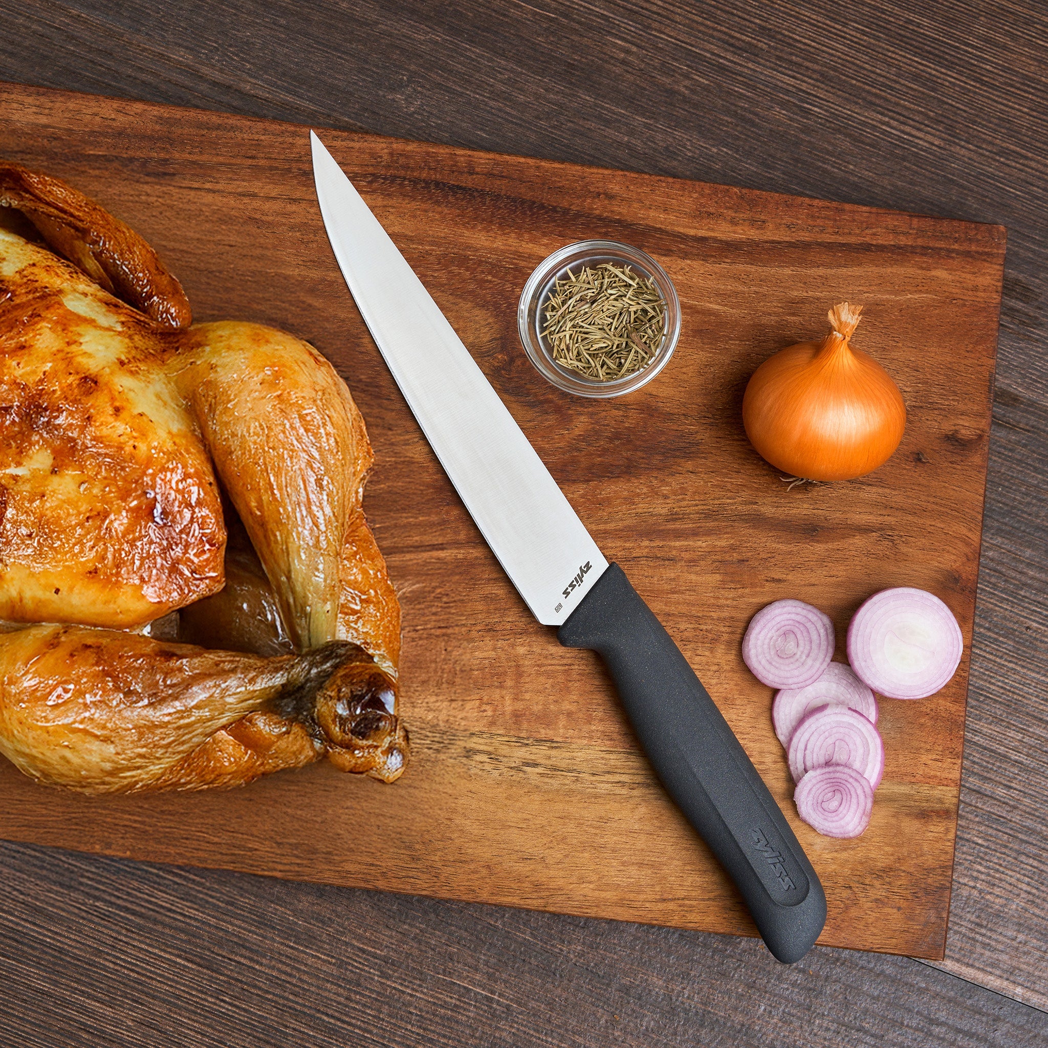 A roasted chicken, Zyliss Large Carving Knife 19cm/7.4" with a sustainable handle, dried herbs in a small bowl, a whole onion, and sliced red onion are arranged on a wooden cutting board.