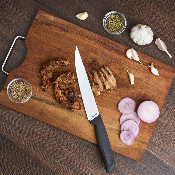 A wooden cutting board on a dark surface holds sliced cooked meat, sliced red onion, rosemary, dried herbs, garlic cloves, and the Zyliss Large Carving Knife 19cm/7.4" with a stainless steel blade and sustainable handle.