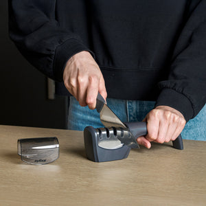 A person maintains kitchen blades by sharpening a knife with the Zyliss Knife Sharpener on a wooden surface, keeping the sharpener cover nearby for safety and cleanliness.