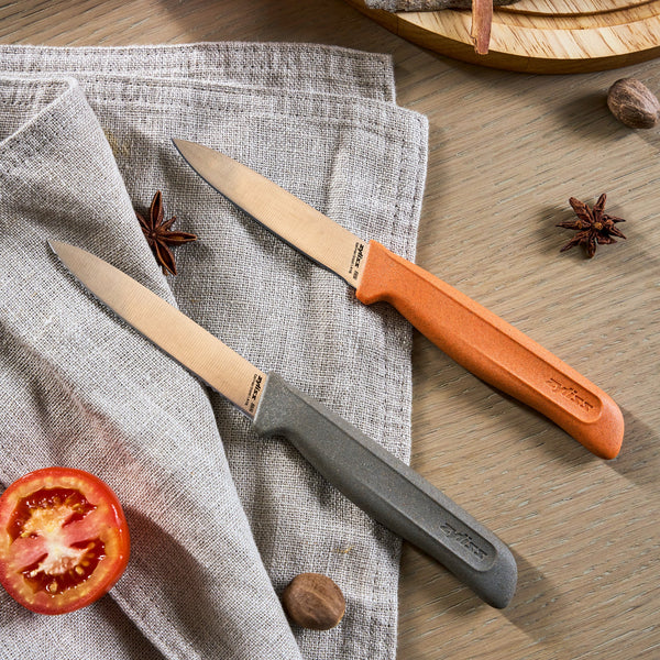 The Zyliss 2 Piece Knife Set, featuring orange and gray handles, rests on a textured beige cloth with star anise, nutmeg, a sliced tomato, and a wooden cutting board.