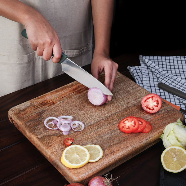 A person uses a Zyliss 3 Piece Knife Set to slice a red onion on a wooden cutting board, surrounded by sliced tomatoes, lemon, lettuce, and a checkered towel—showcasing an organized kitchen setup.