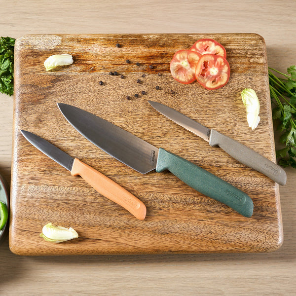The Zyliss 3 Piece Knife Set features vibrant orange, green, and beige-handled knives on a wooden cutting board, surrounded by tomato slices, lettuce, herbs, and scattered peppercorns.