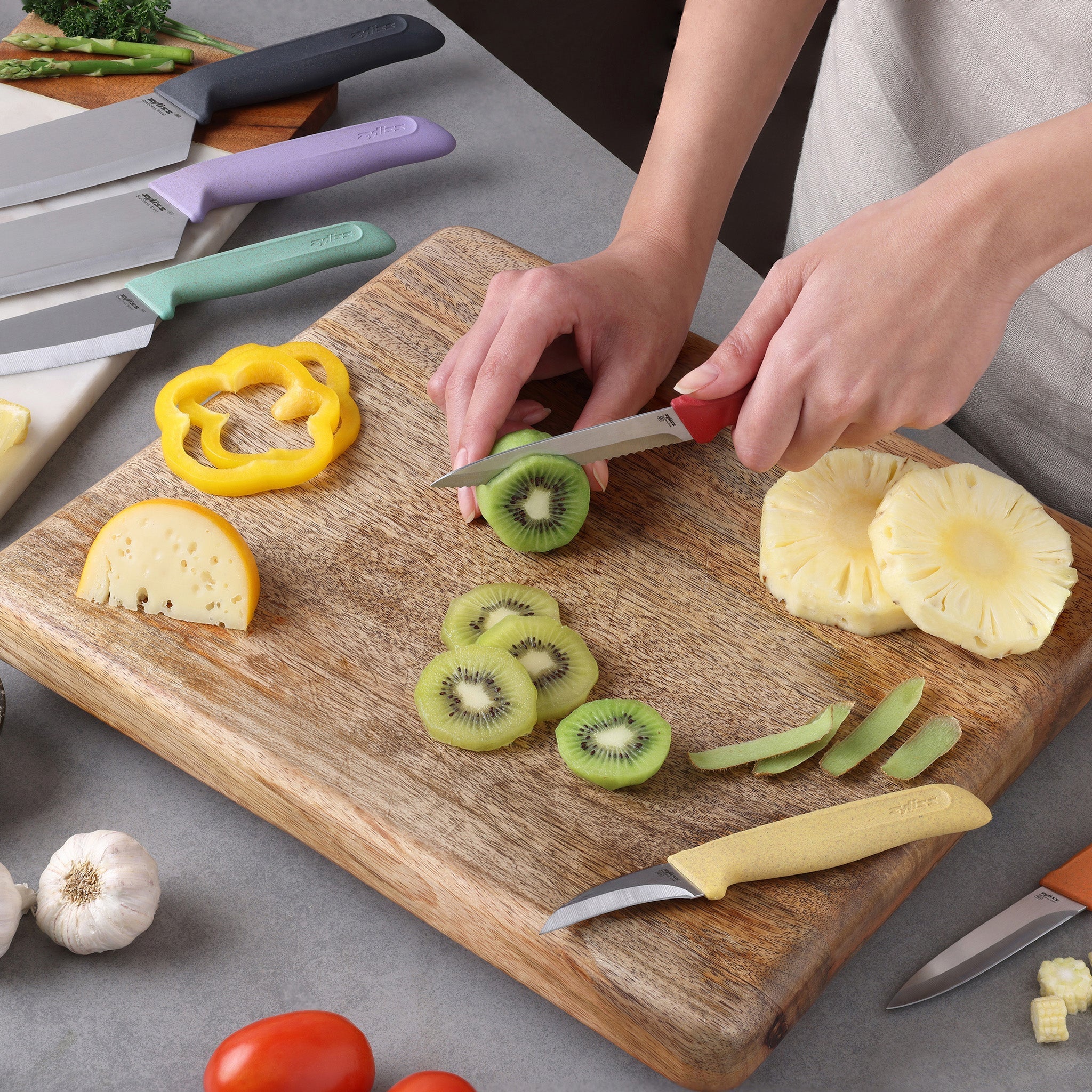 A person uses the Zyliss Classic 6-Piece Knife Set to slice a kiwi on a wooden cutting board. Sliced pineapple, bell pepper, and kiwi are on the board, with garlic, tomato, and herbs nearby on a gray countertop.