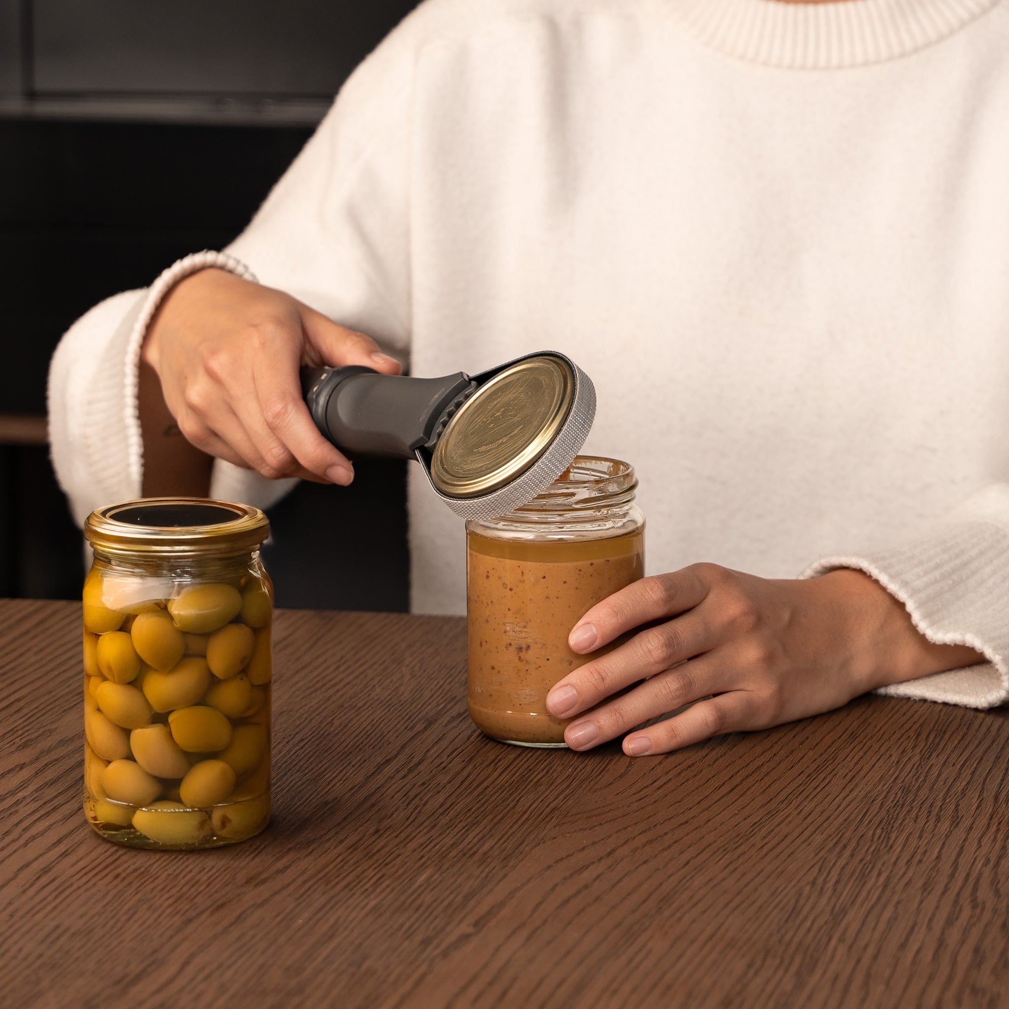 A person uses a jar opener to twist the lid off a glass jar of brown spread, with a sealed jar of green olives nearby on a wooden table.