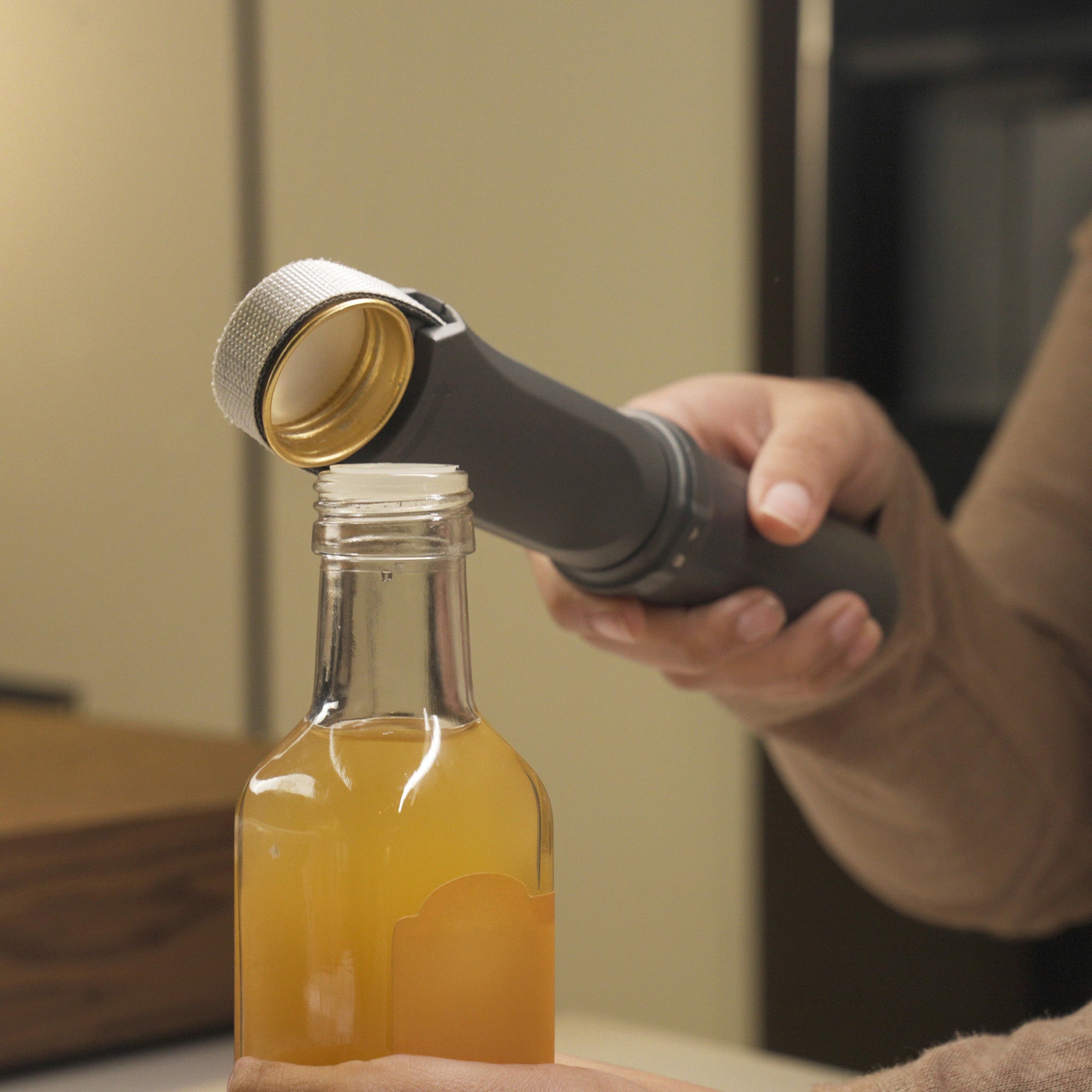 A person uses a metal bottle opener tool to remove the cap from a glass bottle filled with a light orange liquid. The bottle is partially labeled and the scene is set indoors.