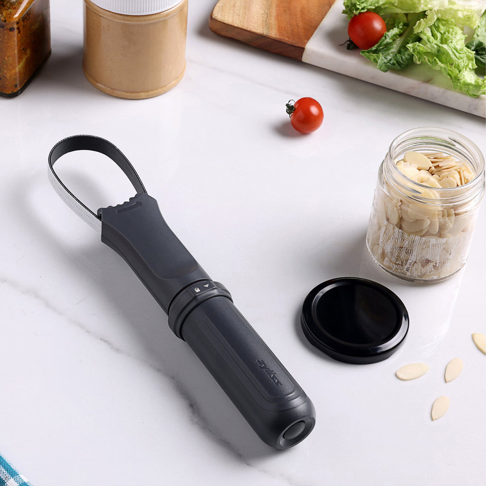 The Zyliss Jar Opener, shown vertically against a light background, is a black and gray handheld tool with a textured grip, integrated locking mechanism, circular top loop, and comes with a protective blade sheath. 