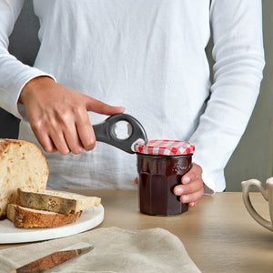A person in a white shirt uses the Zyliss 5 In 1 Opener with an ergonomic grip to open a jar of jam with a red-and-white checkered lid. On the table are slices of bread, a knife, a mug, and a beige napkin.
