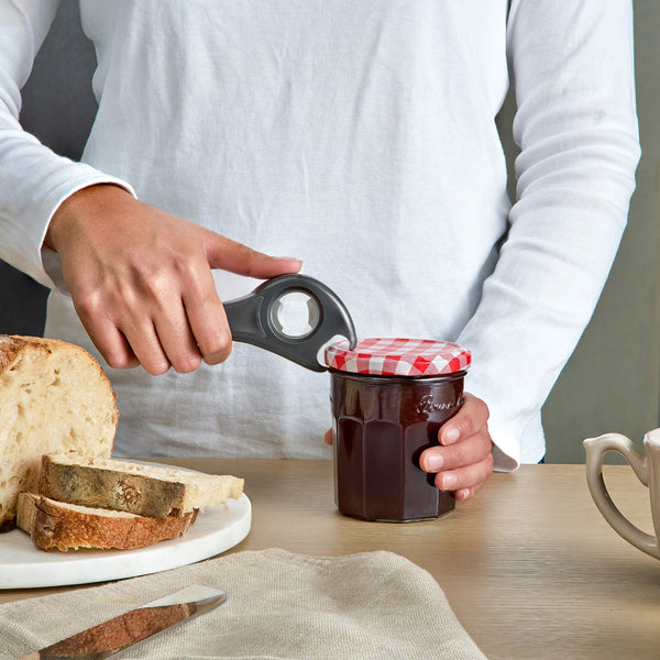 A person in a white shirt uses the Zyliss 5 In 1 Opener with an ergonomic grip to open a jar of jam with a red-and-white checkered lid. On the table are slices of bread, a knife, a mug, and a beige napkin.