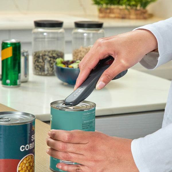 In a kitchen, someone uses the Zyliss 5 In 1 Opener with an ergonomic grip to open a can. On the counter in the background are canned goods, spice jars, a green soda can, and a bowl of salad.