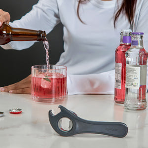 Someone pours a drink from a brown bottle into a glass with pink beverage and ice. Two unopened bottles sit nearby, alongside the Zyliss 5 In 1 Opener with ergonomic grip on the white countertop.