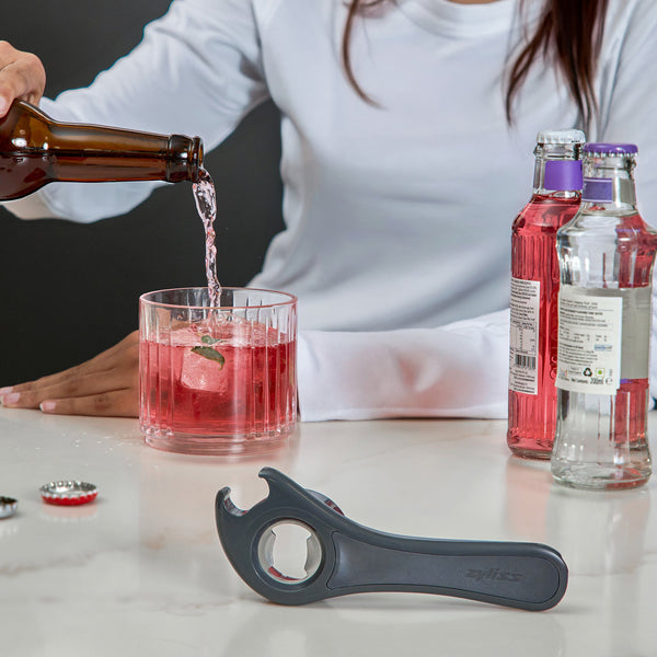 Someone pours a drink from a brown bottle into a glass with pink beverage and ice. Two unopened bottles sit nearby, alongside the Zyliss 5 In 1 Opener with ergonomic grip on the white countertop.