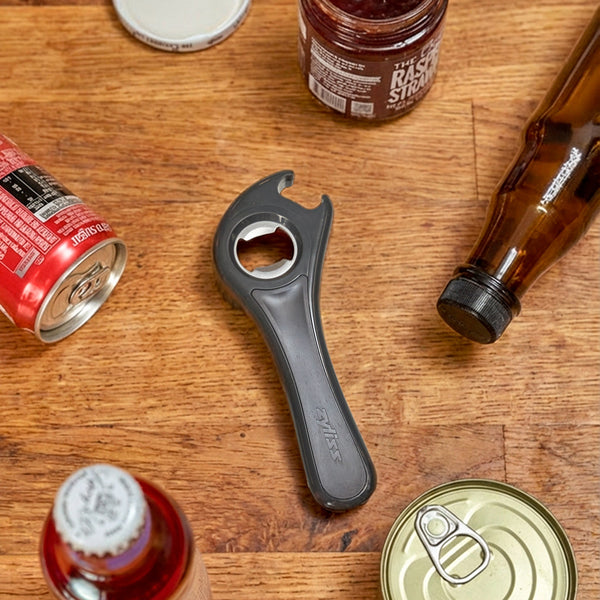 The Zyliss 5 In 1 Opener, a gray compact tool with an ergonomic grip, rests on a wooden surface among assorted bottles and cans, some opened and others still sealed.