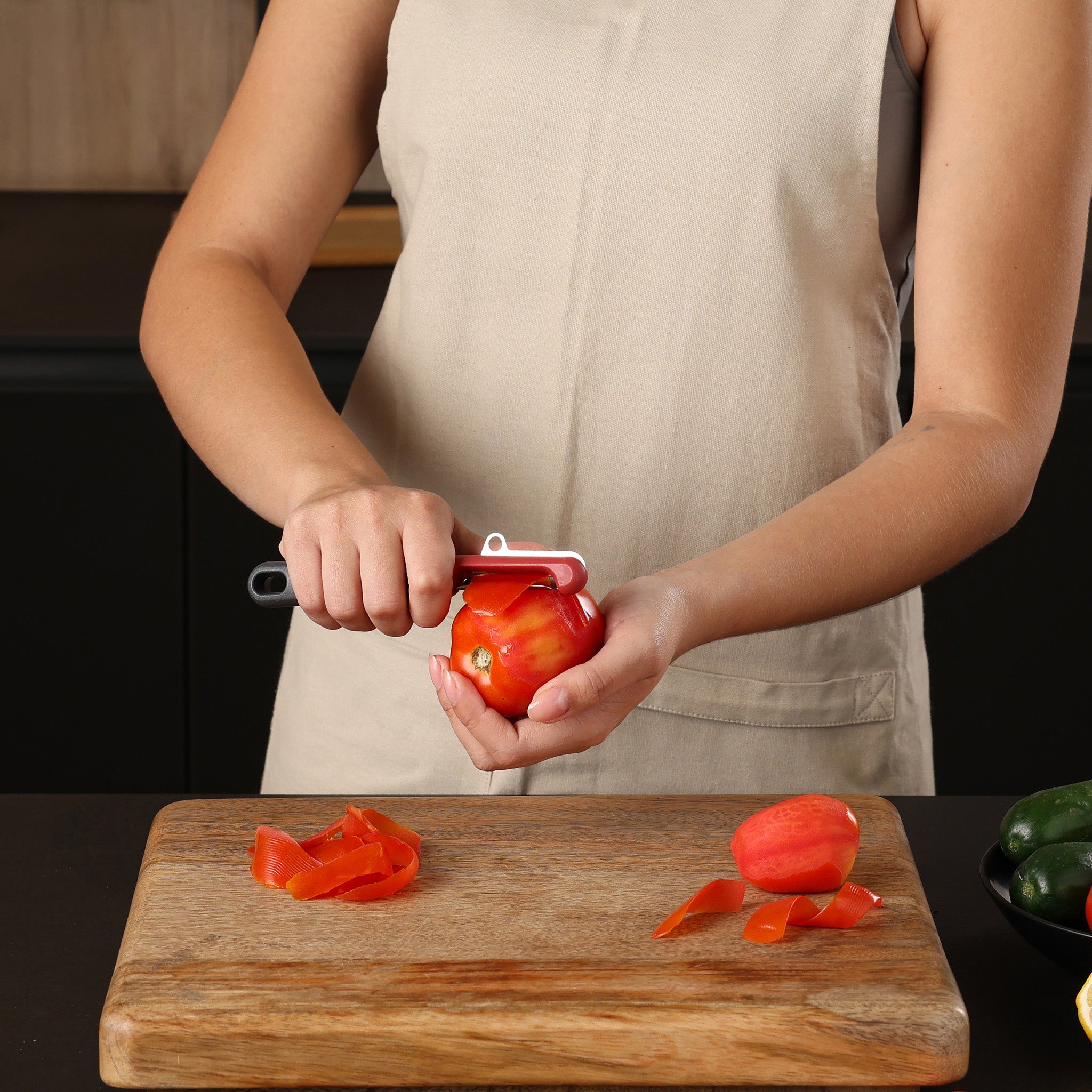 A person wearing a beige apron uses a vegetable peeler to peel the skin off a tomato over a wooden cutting board. Tomato peels and a partially peeled tomato are on the board.