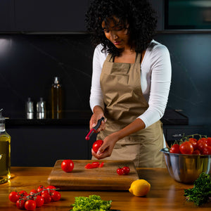 A woman in a beige apron uses the Zyliss Soft Skin Peeler to slice a tomato on a wooden cutting board in a modern kitchen, surrounded by fresh vegetables like tomatoes, lettuce, and a lemon. 