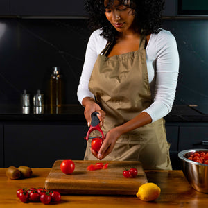 A woman in a beige apron uses the Zyliss Soft Skin Y Peeler to peel a tomato over a wooden cutting board, surrounded by more tomatoes, a lemon, kiwis, and a metal bowl of cherry tomatoes on the kitchen counter. 
