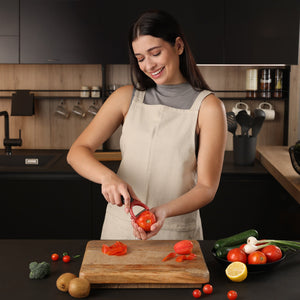 A woman in a beige apron smiles as she peels a tomato with the ergonomic Zyliss Soft Skin Y Peeler in her modern kitchen. A cutting board with a peeled tomato and various fruits and vegetables sits on the counter. 