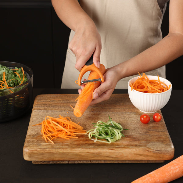 A person uses the Zyliss Julienne Y Peeler to slice a carrot over a wooden cutting board, with shredded carrot, cucumber, cherry tomatoes, greens, and a white bowl nearby—highlighting its ergonomic and eco-friendly design. 