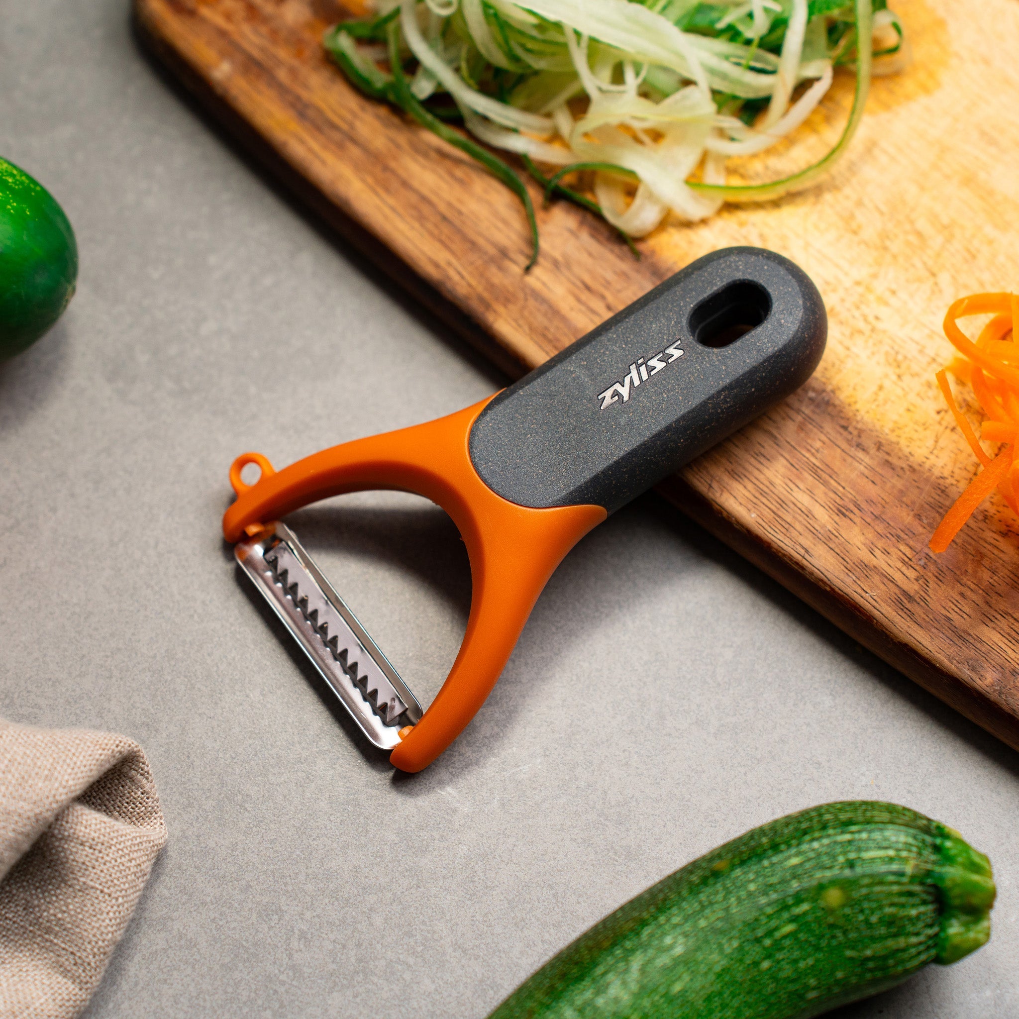 The Zyliss Julienne Y Peeler, featuring an ergonomic orange and black handle, rests on a gray countertop near a wooden board with shredded cucumber and carrot—an essential eco-friendly kitchen tool. 