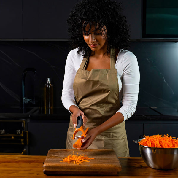 A woman with curly hair, in a beige apron, uses the Zyliss Julienne Y Peeler with an ergonomic grip to shred carrots onto a wooden cutting board in a modern kitchen. A bowl of shredded carrots rests nearby on the counter. 