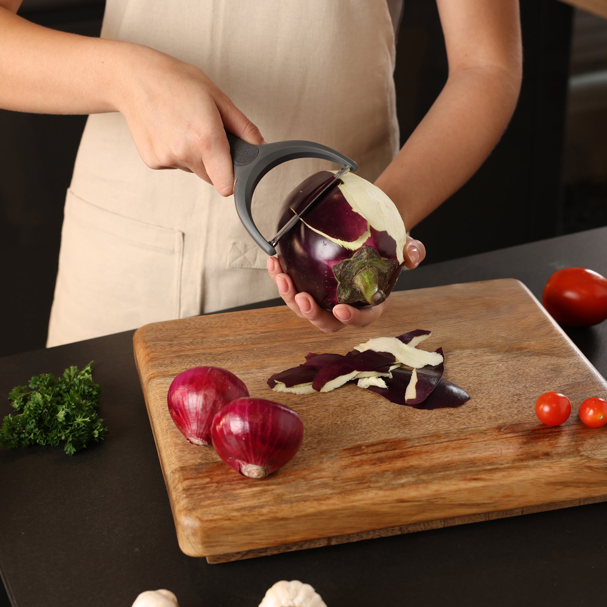 A person in a beige apron is peeling an eggplant with a vegetable peeler over a wooden cutting board. Two red onions, a few cherry tomatoes, fresh herbs, and garlic are also on the board.
