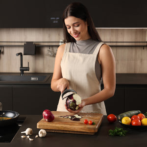 A woman in a modern kitchen smiles as she peels an eggplant with the ergonomic Zyliss Wide Peeler. Fresh tomatoes, zucchini, mushrooms, and parsley sit on the counter beside her. 