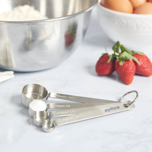 The Zyliss Stainless Steel Measuring Spoons—a kitchen essential—sit on the countertop next to fresh strawberries, eggs, and a stainless mixing bowl filled with flour, perfectly ready for your next baking or cooking task. 