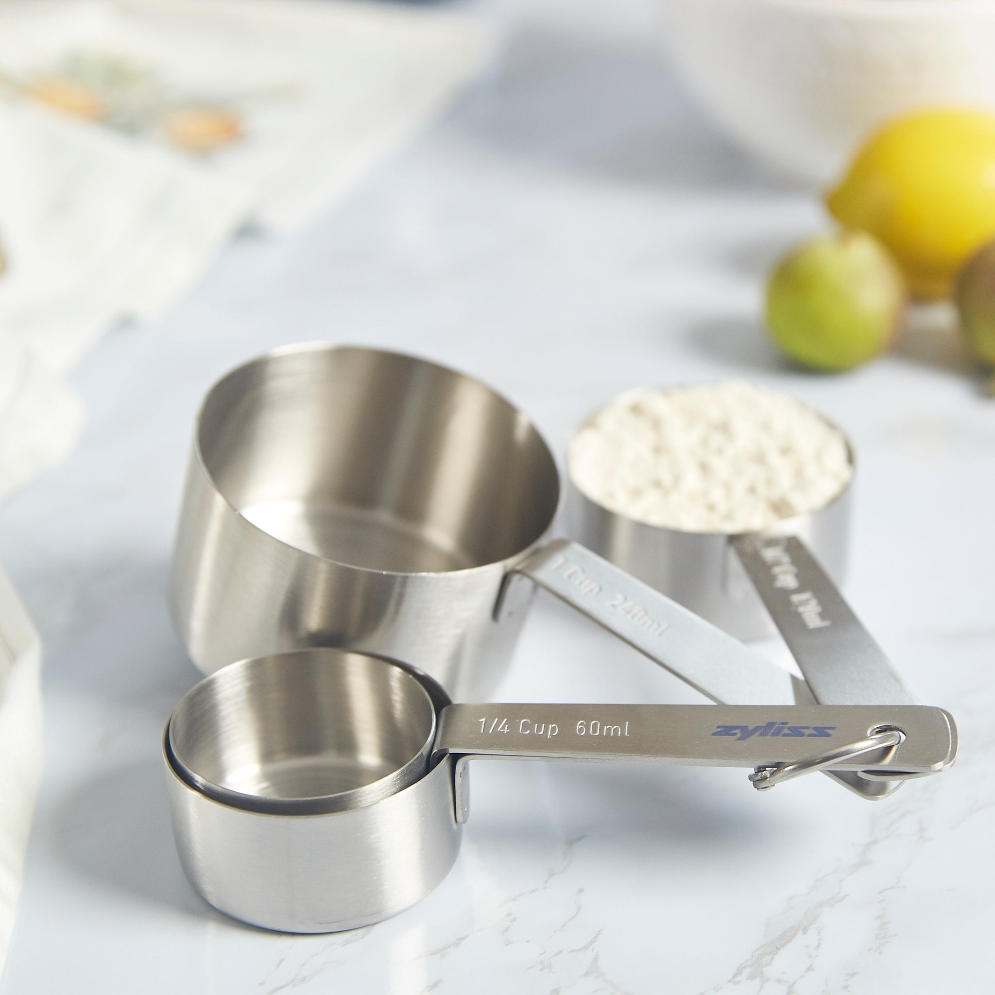 Three Zyliss Stainless Steel Measuring Cups, a must-have for accurate ingredient measuring, are stacked on a marble countertop. Lemons and limes appear blurred in the background. 