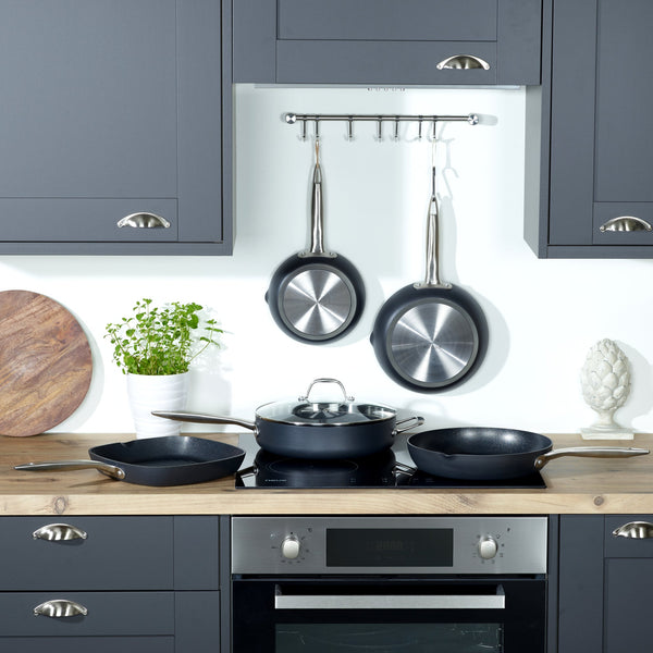 A modern kitchen features dark cabinets, a wooden countertop, and several Zyliss Ultimate Pro Non-Stick Frying Pans with pouring lips—two Swiss-engineered pans hang above the stove, with other cookware and a potted plant on the counter. #20cm/8"