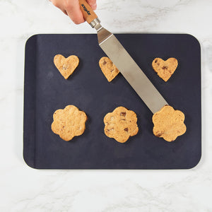 A hand lifts a flower-shaped chocolate chip cookie with a metal spatula from the Zyliss Durable Non-Stick Baking Sheet, while other heart and flower-shaped cookies rest on the sheet atop a white marble surface. 