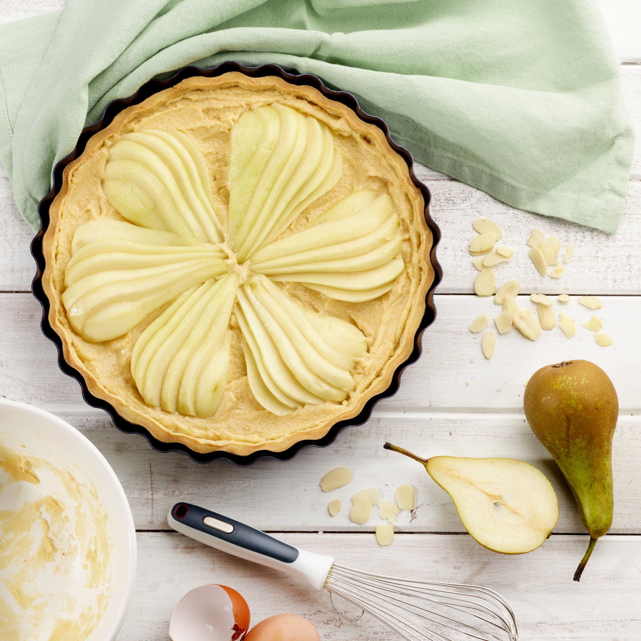 A tart pan filled with unbaked pear tart, arranged with sliced pears in a flower pattern on almond cream. Nearby are a pear, a sliced half, a whisk, eggshells, almond flakes, and a green cloth on a white wooden surface.