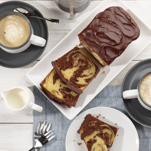 A marble cake with chocolate icing, partially sliced on a white platter, baked in the Zyliss Durable Non-Stick Loaf Pan, is displayed on a white table set with coffee cups, a milk jug, forks, and plates beside a gray napkin. #1.5L/2lb