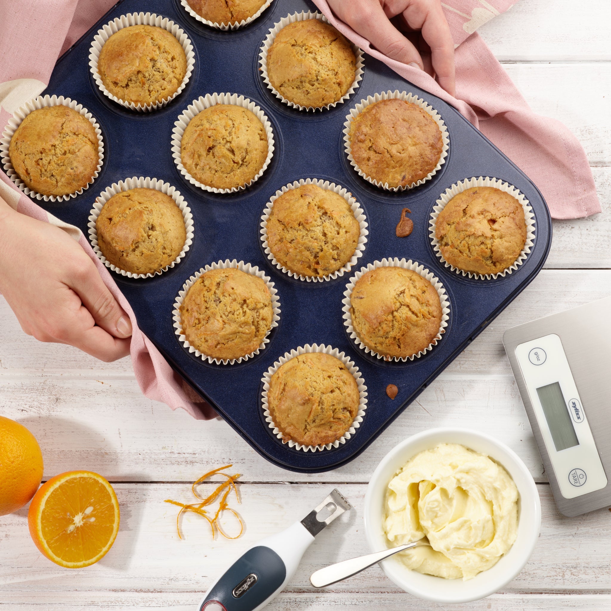 A person holding a muffin tray with twelve baked muffins on a white wooden table, surrounded by a bowl of frosting, a zester, an orange half, orange zest, and a digital kitchen scale.